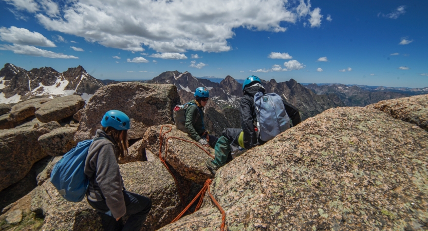 Three people wearing safety gear rest on rocks high above a mountainous landscape.
